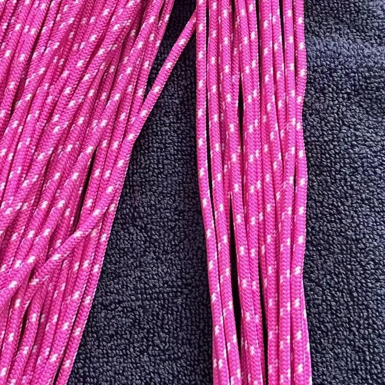 May include: A collection of bright pink paracord strands featuring a repeating white speckled pattern. The cords are arranged in parallel vertical lines, showcasing the braided texture and the vibrant color contrast of the synthetic material.