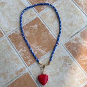May include: A necklace featuring a blue rope cord with small white speckles, connected by gold-toned metal coils and a lobster clasp to a smooth, glossy red heart-shaped pendant that hangs from a series of small circular gold-toned metal rings.