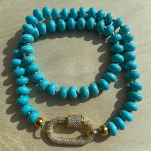 Peut inclure: Un collier de perles turquoise avec un fermoir doré. Le collier est composé de perles ovales enfilées. Le fermoir est de forme ovale dorée, incrusté de petites pierres transparentes. Le collier est présenté sur une surface claire.