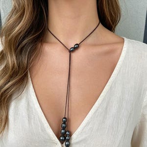 May include: A dark brown lariat necklace with dark grey pearls. The necklace has a simple design with a single strand that splits into two, with pearls clustered at the ends. The necklace is worn against a white top.
