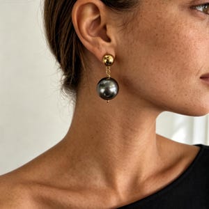 Puede incluir: Un pendiente colgante que presenta una pequeña esfera dorada pulida en el lóbulo, conectada por un eslabón corto dorado a una cuenta esférica más grande, lisa y de color gris oscuro que cuelga debajo. El diseño es sencillo y limpio.