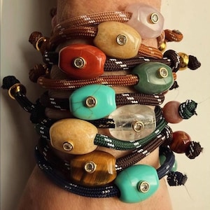 Puede incluir: Una colección de pulseras apiladas con cordones de nailon trenzado en varios colores, cada una adornada con una cuenta de piedra pulida. Cada piedra está engastada con un pequeño detalle circular en tono dorado con una piedra clara.
