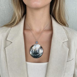 Puede incluir: Un collar con colgante grande y redondo con un diseño de concha natural. El colgante es principalmente negro, blanco y gris, con una cuenta azul claro encima. El collar se lleva sobre una camiseta blanca y una blazer beige.