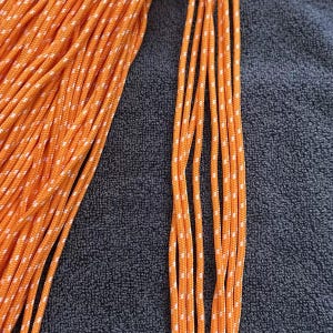 May include: A bundle of bright orange paracord featuring a repeating white reflective fleck pattern woven throughout the length of the cord. The cord is arranged in a dense coil on the left side with several individual strands extending to the right.