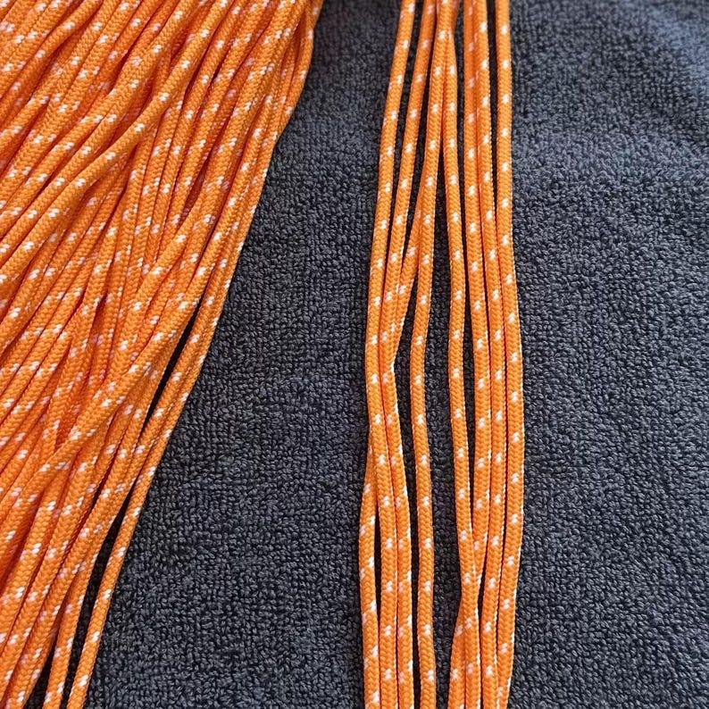 May include: A bundle of bright orange paracord featuring a repeating white reflective fleck pattern woven throughout the length of the cord. The cord is arranged in a dense coil on the left side with several individual strands extending to the right.