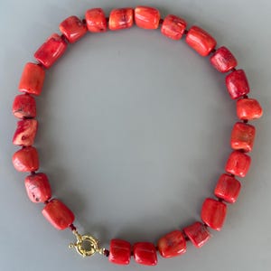 May include: A necklace composed of numerous cylindrical, vibrant red coral beads strung together on a red cord. The necklace features a circular gold-toned metal spring ring clasp at the bottom, which connects the two ends of the beaded strand.