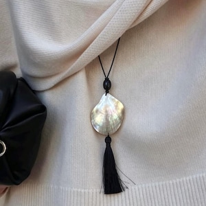 May include: A long necklace featuring a black cord, a single black bead, a large iridescent mother-of-pearl shell pendant, and a black thread tassel hanging at the bottom. The necklace is worn over a cream-coloured knit garment.