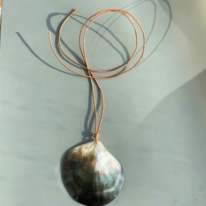 Op de afbeelding: Een ketting met een hanger van een parelmoer schelp en een bruin leren koord. De schelp heeft natuurlijke tinten zwart, grijs en een vleugje groen en blauw. Het koord is bovenaan geknoopt, wat een eenvoudig, elegant ontwerp cre&euml;ert.