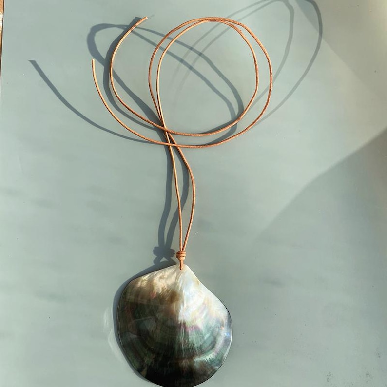 Op de afbeelding: Een ketting met een hanger van een parelmoer schelp en een bruin leren koord. De schelp heeft natuurlijke tinten zwart, grijs en een vleugje groen en blauw. Het koord is bovenaan geknoopt, wat een eenvoudig, elegant ontwerp cre&euml;ert.