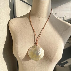 Op de afbeelding: Een ketting met een grote, parelmoer schelpenhanger. De schelp is cr&egrave;mewit met iriserende highlights, hangend aan een dun bruin leren koord. De ketting wordt getoond op een mannequin, die de natuurlijke schoonheid van de schelp benadrukt.