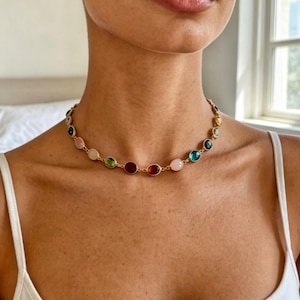 May include: A gold-toned chain necklace featuring a series of evenly spaced, round, multi-coloured cabochon stones. The stones display a variety of hues including pink, green, red, orange, and blue, set in individual circular metal bezels.