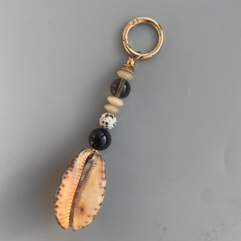 May include: A seashell keychain with a gold-colored clasp. The keychain features a string of beads in various colors, including black, white, and tan. The seashell is a light brown color with darker brown markings.