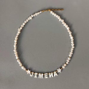 May include: A white pearl necklace with the name "JIMENA" spelled out in white letters. The necklace has gold-colored accents and star-shaped charms on each side of the name. The necklace is displayed on a gray background.