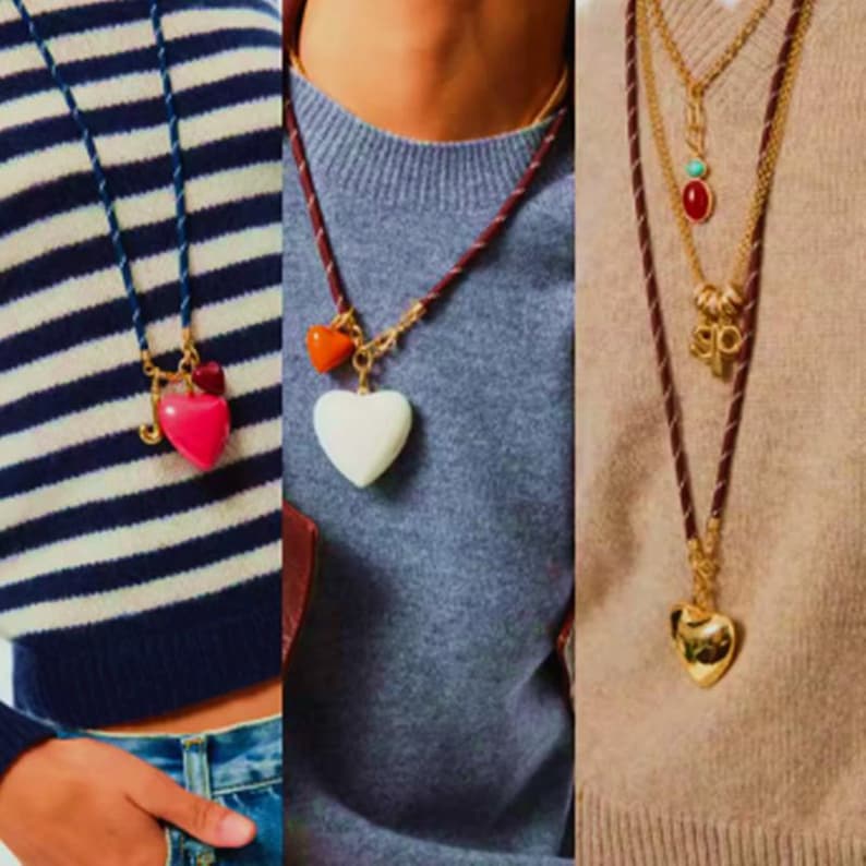 May include: Three necklaces featuring heart-shaped pendants. The first is a blue cord with a pink heart, the second a brown cord with a white heart and orange accent, and the third a brown cord with a gold heart and a multi-layered gold chain.