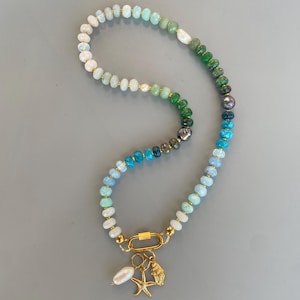 Puede incluir: Un collar de cuentas con un degradado de piedras preciosas redondas y facetadas en tonos blanco, azul claro, turquesa, verde y gris oscuro. El collar incluye un cierre de mosquetón ovalado en tono dorado con dijes de estrella y concha.