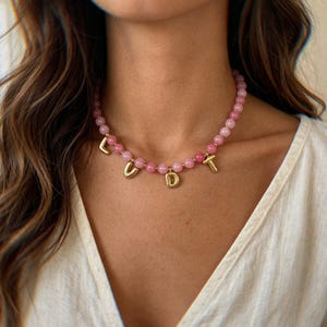 Pode incluir: Um colar de contas composto por contas redondas de pedra preciosa rosa claro enfiadas. Entre as contas, quatro berloques metálicos dourados em forma de letra formam a palavra LUDT, pendurados como pingentes na corrente circular de contas.