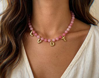 May include: A beaded necklace featuring round, light pink gemstone beads strung together. Interspersed among the beads are four gold-toned, metallic letter charms that spell out the word LUDT, hanging as pendants from the circular beaded chain.