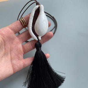 Puede incluir: Un collar con colgante de cerámica blanca con una borla negra. El colgante tiene forma de lágrima y está sujeto a un cordón marrón. El collar se sostiene en una mano.