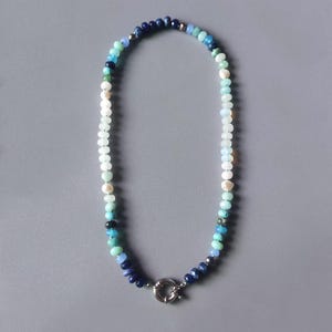 May include: A beaded necklace featuring a variety of blue, green, and white beads. The necklace has a silver-colored clasp and is displayed against a gray background. The beads vary in size and shade, creating a gradient effect.
