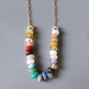 May include: A necklace with a gold chain and a strand of polished, multi-coloured gemstone beads. The beads are arranged in a gradient of colours, including yellow, orange, red, pink, blue, and green. The necklace is displayed against a grey background.