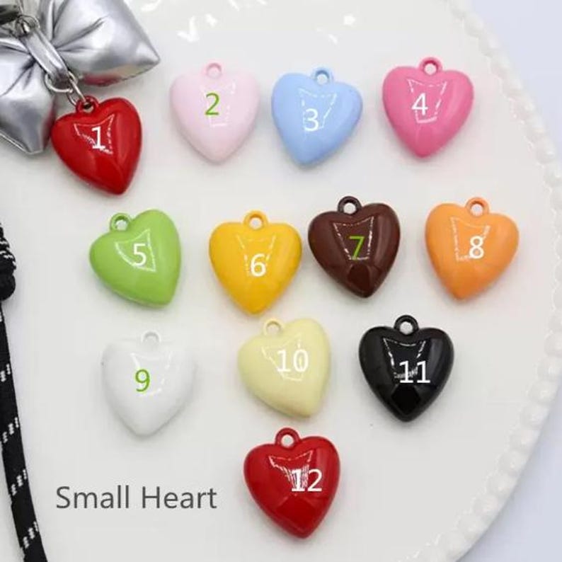 May include: A collection of twelve heart-shaped charms in various colors, each labeled with a white number from 1 to 12. The charms are glossy and smooth, featuring a small loop at the top for attachment. The colors include red, pink, blue, green, yellow, brown, orange, white, cream, and black.