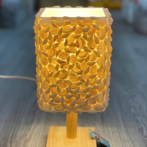 Può includere: Lampada da tavolo rettangolare con paralume ricoperto di piccole conchiglie chiare. La lampada ha una base e un supporto in legno. La lampada emette una luce gialla calda, creando un'atmosfera accogliente. Ideale per camera da letto o soggiorno.
