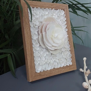Peut inclure: Une œuvre d'art encadrée avec un motif floral central fait de coquillages blancs, entouré de petits coquillages. L'œuvre est encadrée dans un cadre en bois naturel, créant une esthétique côtière ou sur le thème de la plage.
