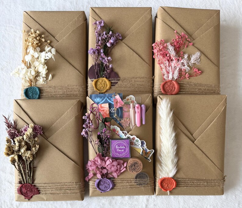 May include: Six rectangular packages wrapped in brown paper, each adorned with dried flowers, twine, and wax seals in various colors. Some packages feature stickers and decorative elements. The overall aesthetic is rustic and artisanal.