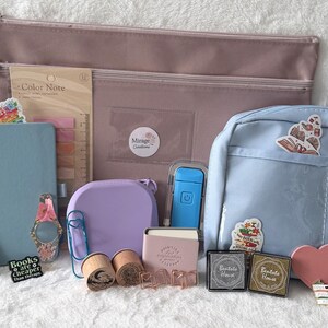 May include: An assortment of stationery items, including a light blue notebook, pens, stickers, and a pink pencil case. Other items include a purple container, a small pink notebook, and a blue backpack-style pouch. The items are arranged on a white surface.