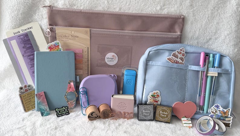 May include: An assortment of stationery items, including a light blue notebook, pens, stickers, and a pink pencil case. Other items include a purple container, a small pink notebook, and a blue backpack-style pouch. The items are arranged on a white surface.