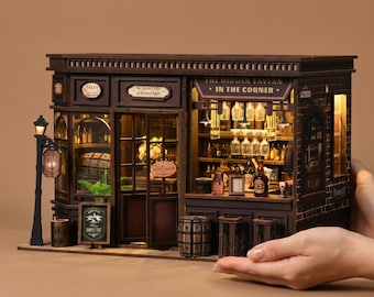 May include: A miniature wooden tavern model with intricate details, including a sign that reads "The Hidden Tavern - In The Corner." The model features a bar, shelves of bottles, and a street lamp. The model is held in a person's hands.