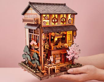 May include: A miniature wooden Japanese restaurant model with intricate details, including a shingled roof, paper lanterns, and a small cherry blossom tree. The model is held in someone's hands against a pink background.