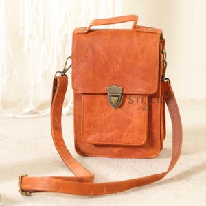 Può includere: Borsa a tracolla rettangolare in pelle marrone-arancio con manico superiore e tasca frontale. La borsa ha una chiusura con fermaglio in metallo e una lunga tracolla regolabile. Il testo "STITCH & CAR" è visibile.