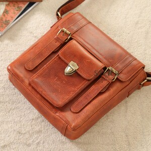 May include: A brown leather messenger bag with a front pocket secured by a metal clasp. The bag features adjustable straps and brass-colored buckles. The leather has a textured appearance.