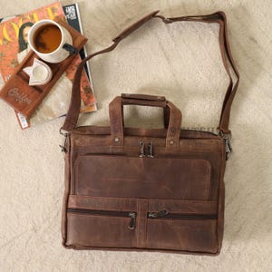 Op de afbeelding: Een bruine leren messenger bag met een handvat aan de bovenkant en een schouderriem. De tas heeft meerdere vakken met ritssluiting en een voorvak. Een houten dienblad met een kopje koffie en een tijdschrift staan op de achtergrond.