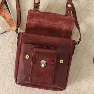 May include: A burgundy leather crossbody bag with a flap and a front pocket. The bag has a shoulder strap and gold-colored hardware. The interior is visible, revealing a suede-like lining. The bag is open, showcasing its storage capacity.