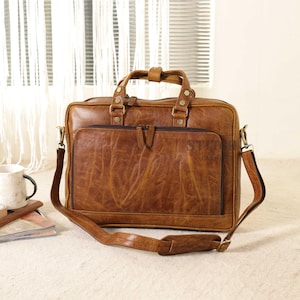 May include: A brown leather briefcase with a top handle and a detachable shoulder strap. The bag has a front pocket with a zipper and a flap. The leather has a distressed look. The bag is approximately 16 inches wide and 12 inches tall.