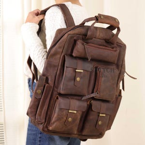 Peut inclure: Un sac à dos en cuir marron avec plusieurs poches extérieures et une poignée supérieure. Le sac à dos a une finition vieillie et comporte un compartiment principal zippé et plusieurs petites poches avec fermetures à rabat. Le texte "ITCH & BRRY" est visible.