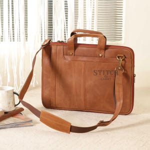 May include: A brown leather briefcase with a handle and a detachable shoulder strap. The bag has a front pocket and the text "STITCH & CARRY" printed on the front. The interior is lined with red fabric.