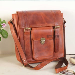 May include: A brown leather messenger bag with a flap closure, front pocket, and adjustable shoulder strap. The bag features brass-colored buckles and hardware. The bag has the text "STITCH" on the front pocket.