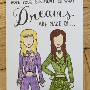 May include: A birthday card with a handwritten message that says "Hope your birthday is what dreams are made of..."  The card features two cartoon women with glitter accents on their clothing. One woman is wearing a purple outfit and the other is wearing a green outfit.