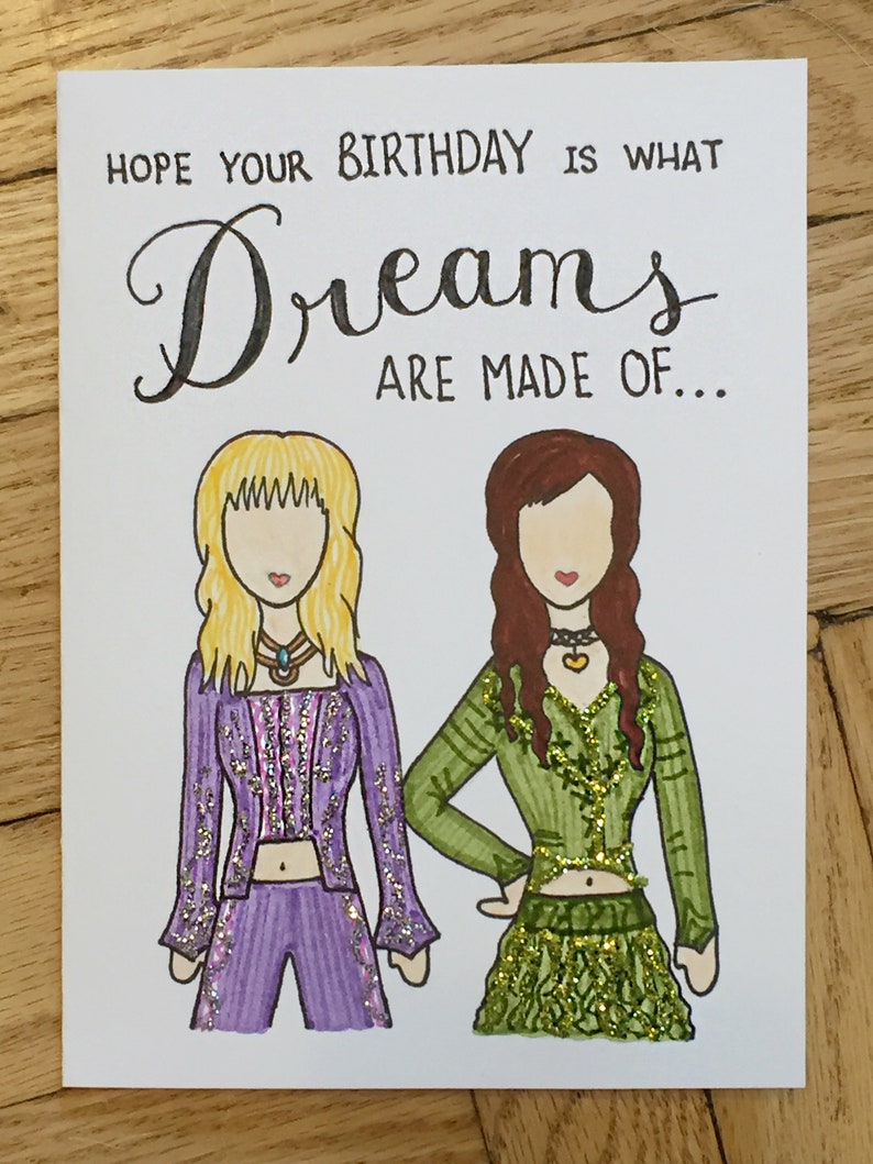 May include: A birthday card with a handwritten message that says "Hope your birthday is what dreams are made of..."  The card features two cartoon women with glitter accents on their clothing. One woman is wearing a purple outfit and the other is wearing a green outfit.