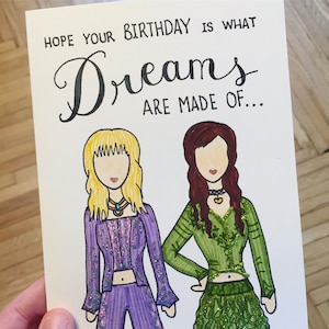 May include: A birthday card with a handwritten message that reads "Hope your birthday is what dreams are made of..."  Two cartoon figures are drawn on the card. One figure is wearing a purple outfit with glitter accents and the other figure is wearing a green outfit with glitter accents.