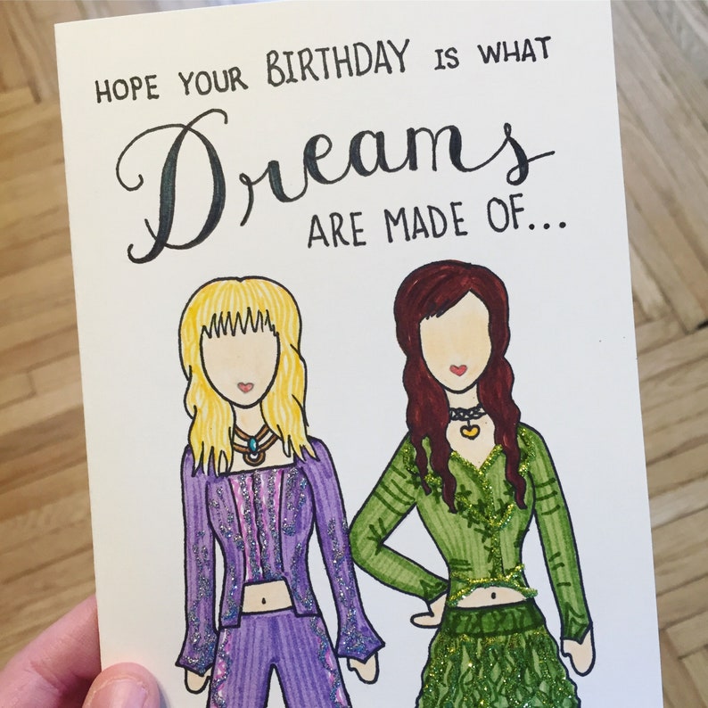May include: A birthday card with a handwritten message that reads "Hope your birthday is what dreams are made of..."  Two cartoon figures are drawn on the card. One figure is wearing a purple outfit with glitter accents and the other figure is wearing a green outfit with glitter accents.