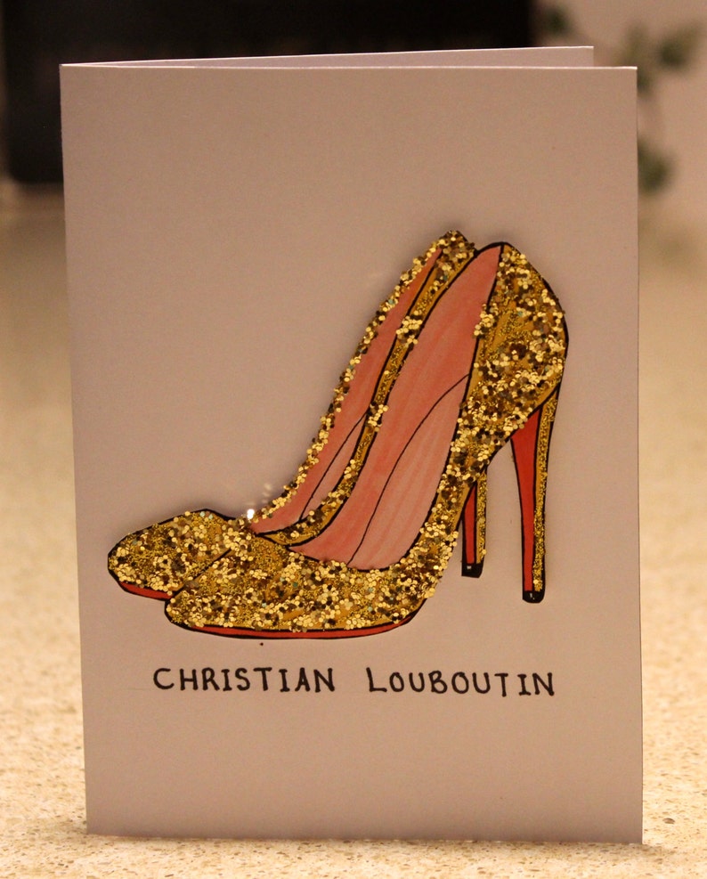 May include: A white card with a drawing of two gold glitter high heel shoes with red soles. The text "CHRISTIAN LOUBOUTIN" is printed below the shoes.