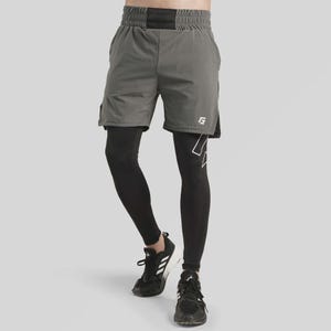 Puede incluir: Pantalones cortos deportivos grises con cinturilla negra y detalle de logotipo blanco. Los pantalones cortos están superpuestos a mallas negras. Zapatillas deportivas negras con detalles blancos completan el conjunto. La imagen está sobre un fondo gris claro.
