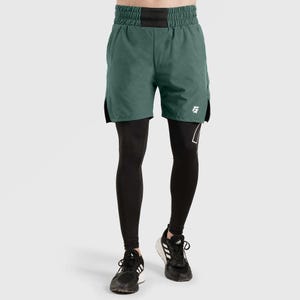 Puede incluir: Pantalones cortos deportivos verdes con cintura negra y aberturas laterales, superpuestos a mallas de compresión negras. Los pantalones cortos tienen un logotipo blanco en la pierna. Zapatillas deportivas negras con rayas blancas completan el conjunto.