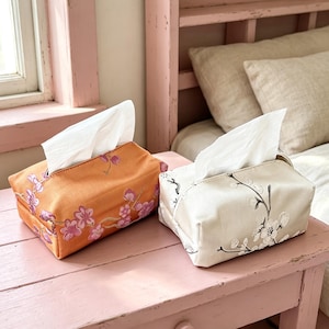 Pode incluir: Duas capas retangulares de tecido para caixas de lenços. Uma capa é laranja com um padrão de flores de cerejeira rosa, e a outra é creme com um bordado de flores de cerejeira brancas e pretas. Ambas contêm lenços brancos na abertura superior.