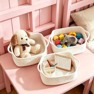 May include: Three white woven baskets on a pink table. One basket holds a stuffed puppy with a brown bow. Another contains colorful wooden blocks and a gray elephant toy. The third basket has pencils and a small calendar.