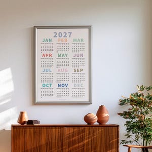 May include: A framed 2027 wall calendar featuring a grid layout of all twelve months. Each month name is printed in a distinct pastel color above its corresponding numerical date grid. The calendar is displayed in a simple silver frame.
